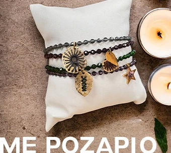 Χειροποίητα βραχιόλια με χάντρες και χρυσά μοτίφ, τοποθετημένα σε μαξιλαράκι, δίπλα σε διακοσμητικά κεριά.