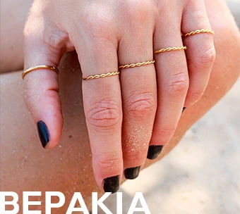 Χέρι με κομψά χρυσά δαχτυλίδια σε minimal σχέδιο, φορεμένα σε διαφορετικά δάχτυλα, με φόντο αμμουδιά.